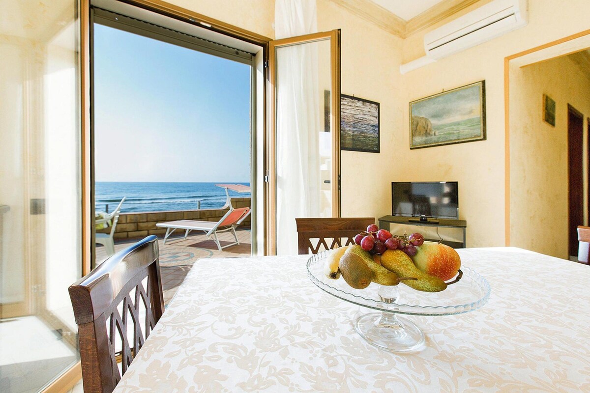 Un comedor con una mesa preparada para dos personas, un frutero sobre la mesa, un televisor en un mueble bajo y vistas al mar a través de una puerta abierta.