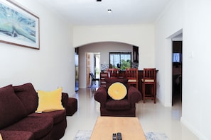 A living room with a sofa, armchair, and coffee table, a dining area with chairs and a table, and a hallway leading to a staircase.
