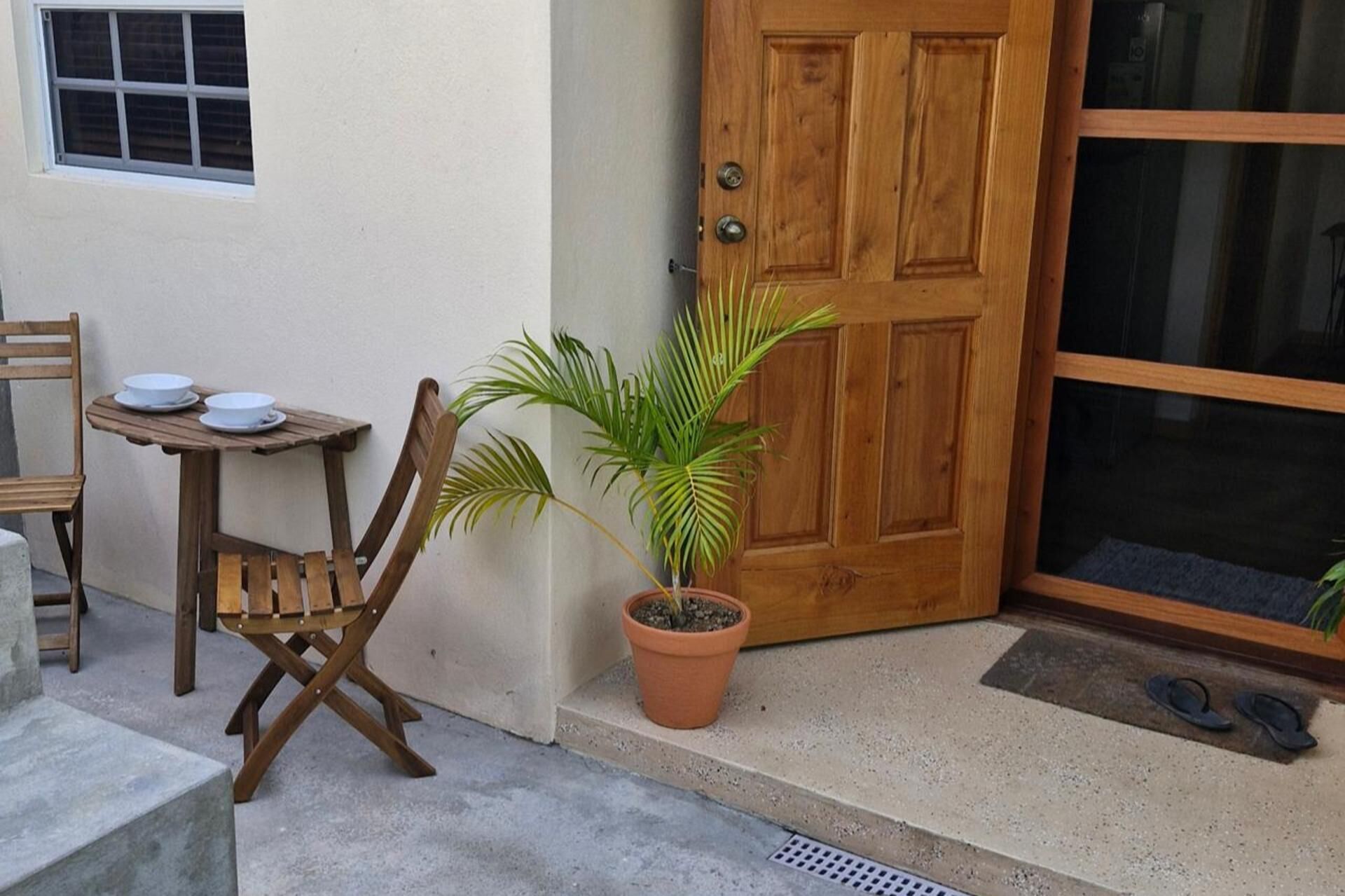 Une porte en bois, une plante en pot, une table avec deux bols, et une chaise devant un bâtiment.