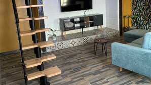 Un salón moderno con una escalera espiral de madera, un televisor de pantalla plana montado en una pared de ladrillo blanco, una chimenea con azulejos estampados y un sofá azul.