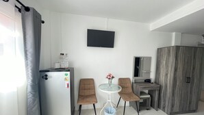 A compact room with a refrigerator, a flat-screen TV, two chairs, a small table with a vase, a cabinet, and a trash bin. - Royal BJ Hotel (Pattaya)