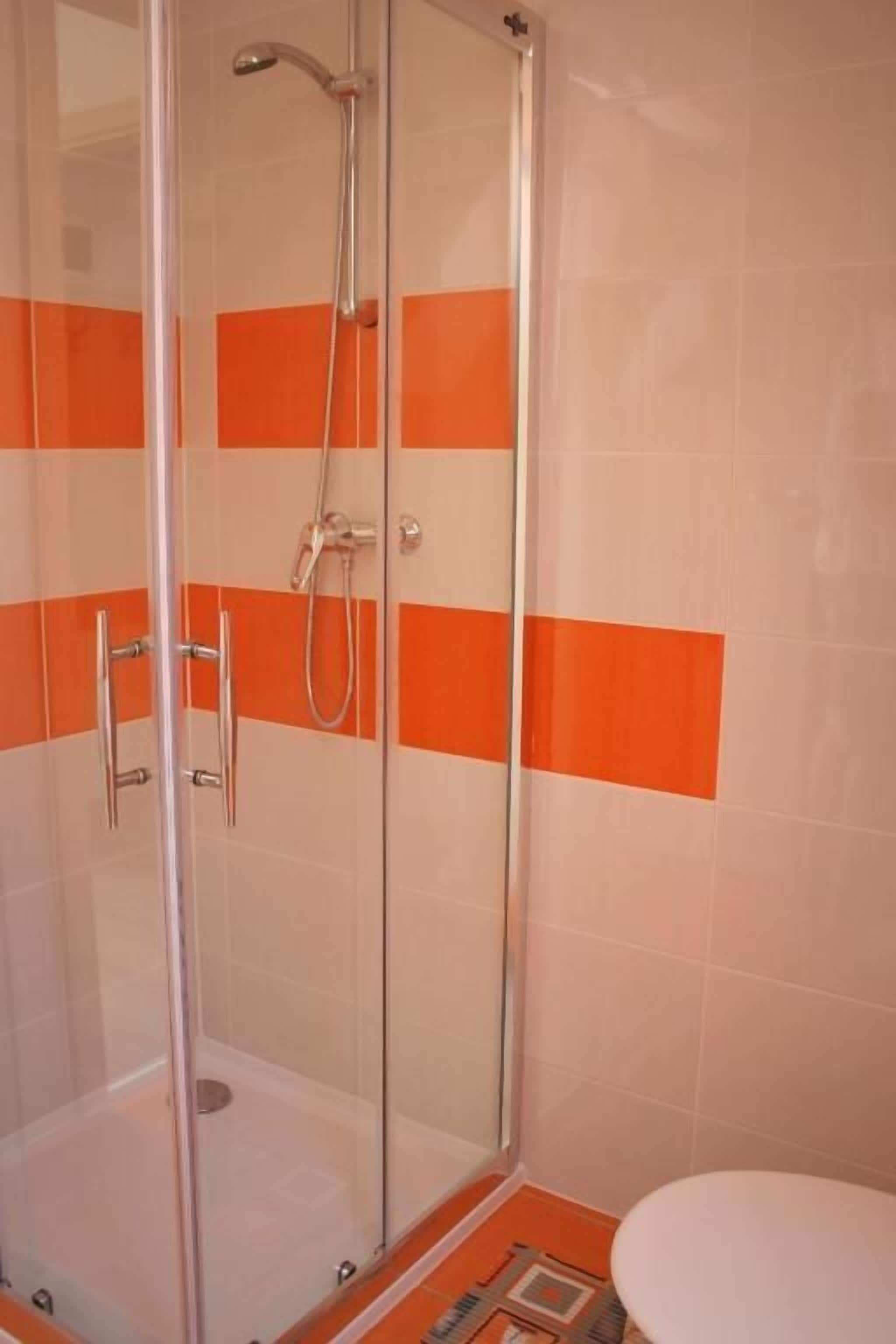 A shower with a glass door, a white toilet, and orange and white tiled walls.