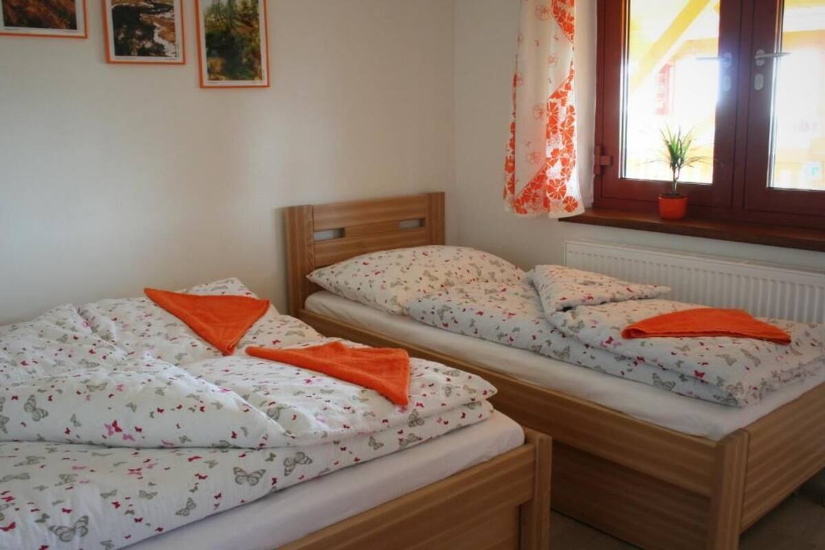 A bedroom with two single beds, each with a wooden headboard, white bedding with a butterfly pattern, and orange throw pillows. There is a window with a curtain and a potted plant on the windowsill.
