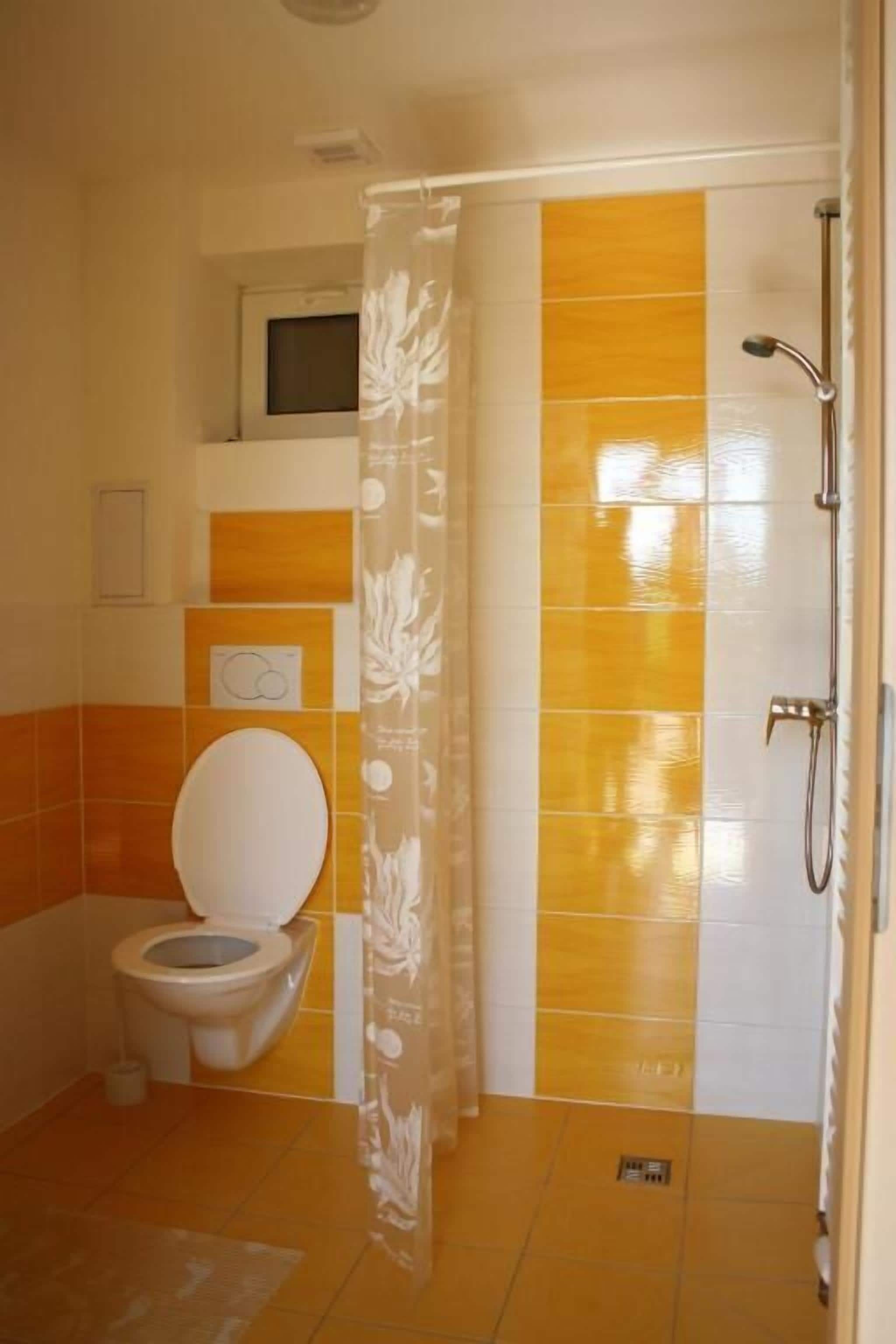 Une salle de bain comprenant des toilettes, une douche et un mur recouvert de carreaux aux couleurs jaune et blanc.