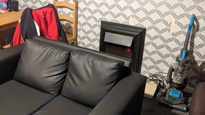 Ein Wohnzimmer mit einem schwarzen Ledersofa, einem Holzstuhl, einem kleinen Tisch mit einer roten Jacke und einem Staubsauger.