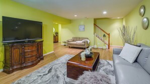 A living room with a flat-screen TV on a wooden cabinet, a gray sofa, a coffee table, and a staircase in the background.