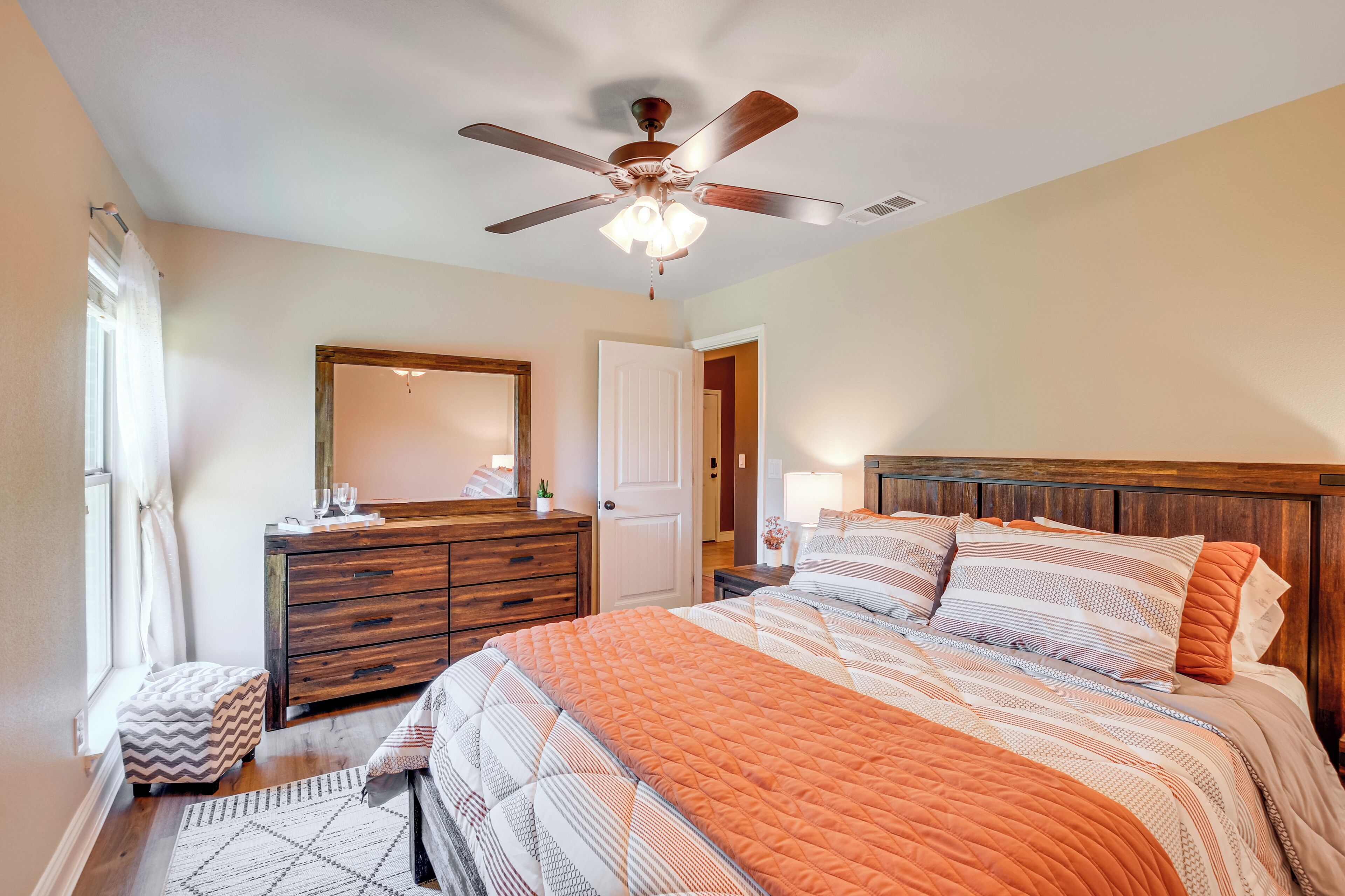 Une chambre à coucher avec une commode en bois, un lit avec une literie à motifs, un ventilateur de plafond et une fenêtre avec des rideaux.