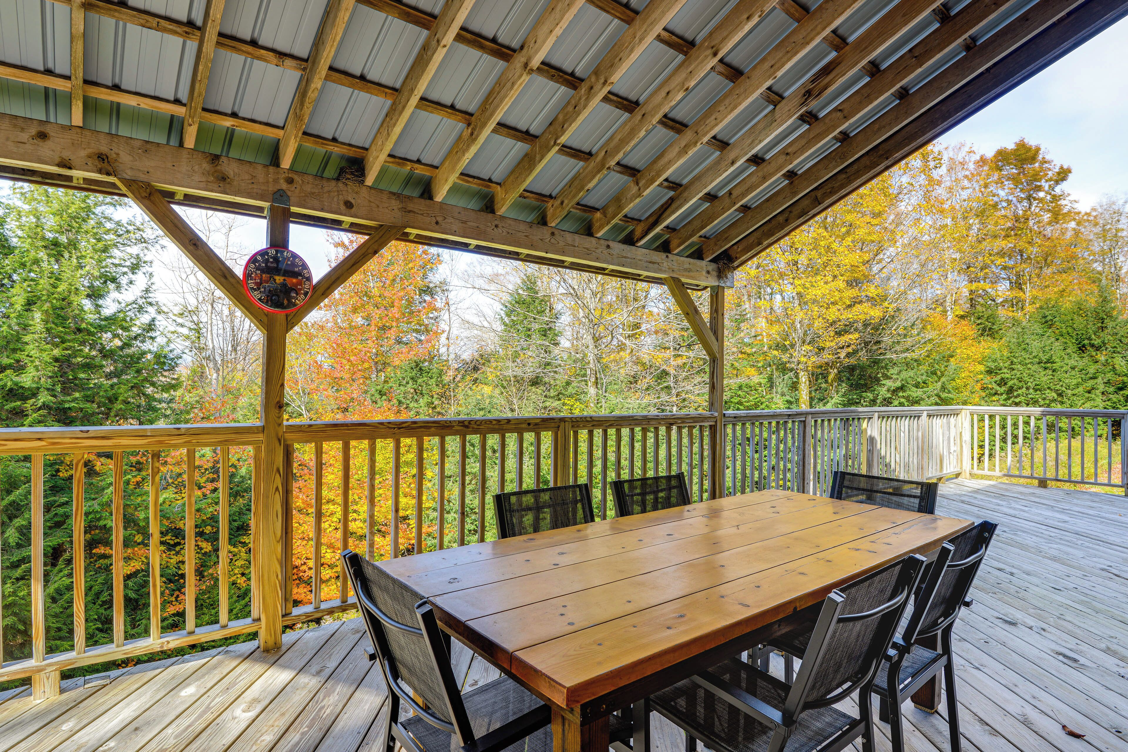 A wooden deck with a table and chairs, surrounded by a wooden railing, overlooking a forest with autumn foliage.