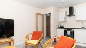 Un espacio habitable compacto con una televisión, dos sillas de mimbre con cojines, una pequeña mesa, una kitchenette con estufa y microondas, y una puerta que da a otra habitación.