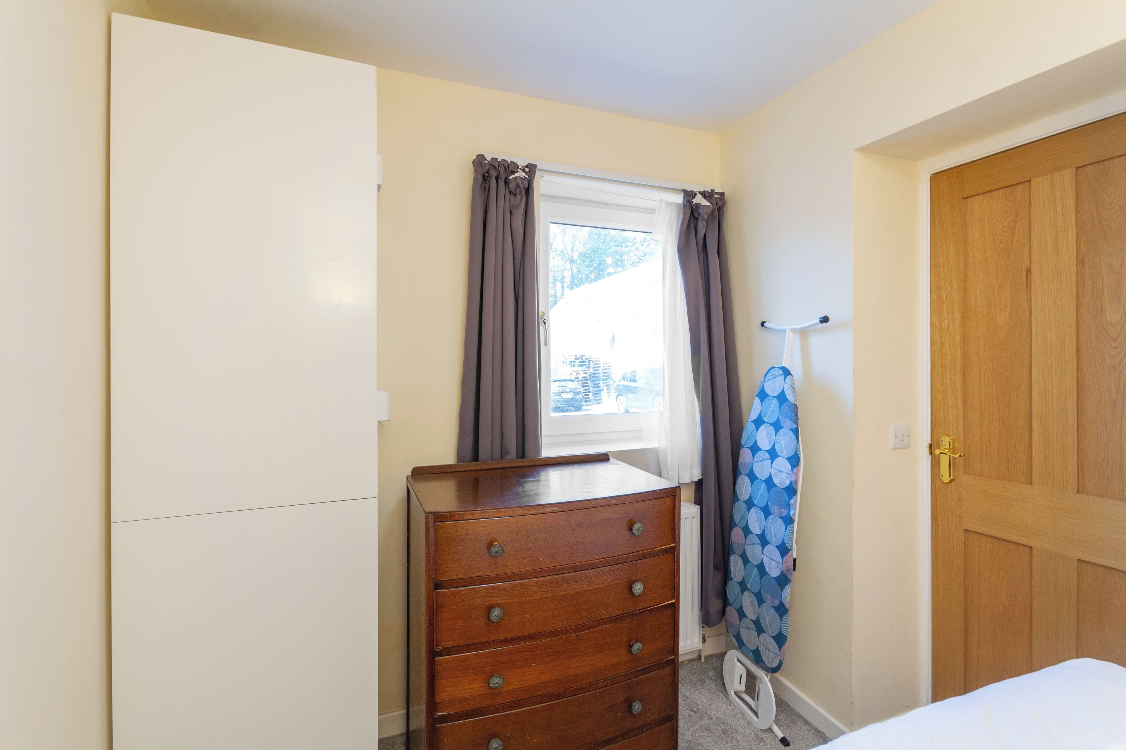 Um quarto com cômoda de madeira, janela com cortinas, porta e cama.