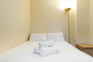 A neatly made bed with white linens and two rolled towels, a bedside table, and a floor lamp.