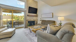 A living room with a sofa, armchair, and coffee table. There is a fireplace, a TV mounted on a wooden wall, and a balcony with outdoor furniture. - 7 Mi to Dwtn - Family Condo in Harbor Springs! (Harbor Springs)