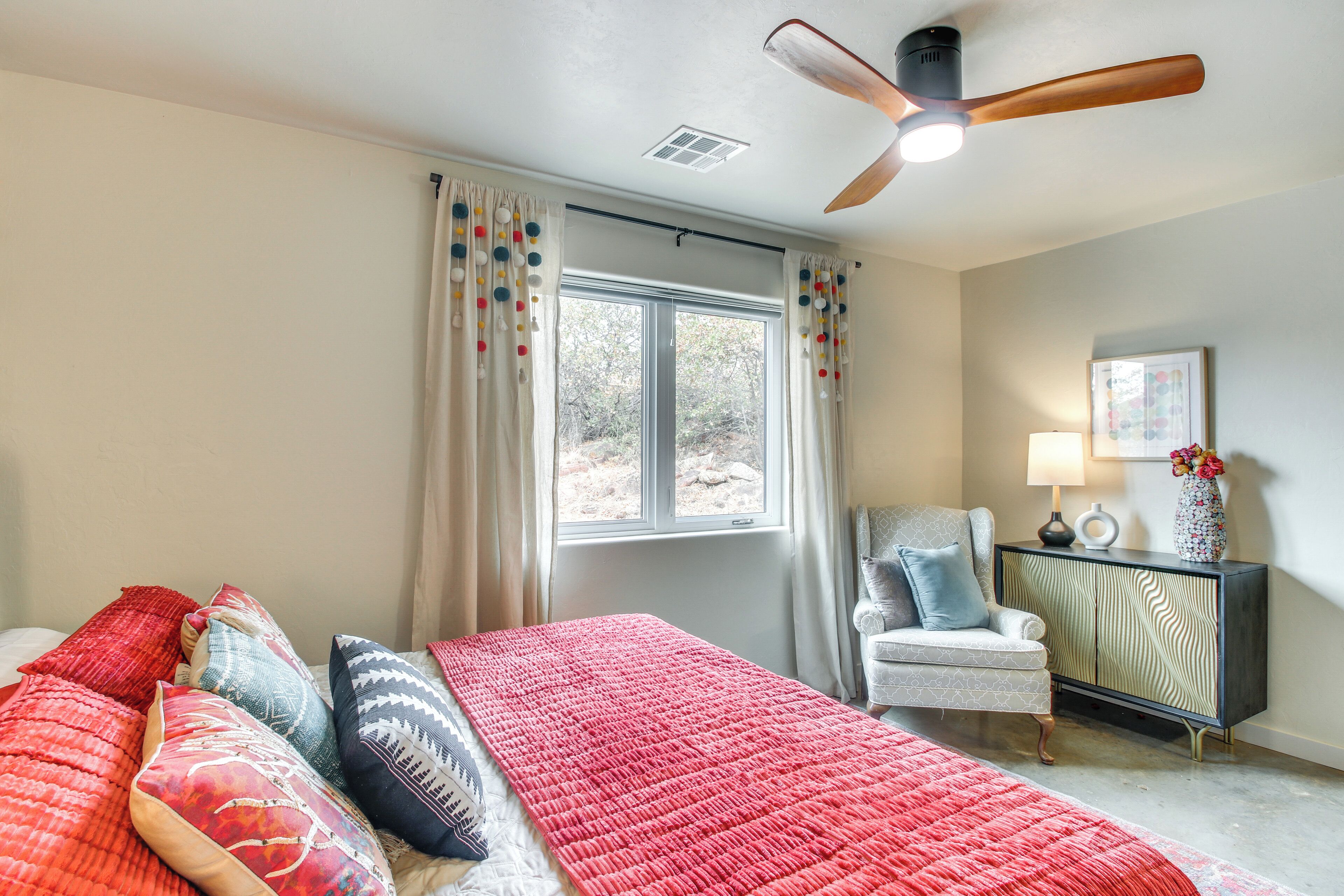 A bedroom with a large bed, a ceiling fan, a window with curtains, a chair, and a cabinet.