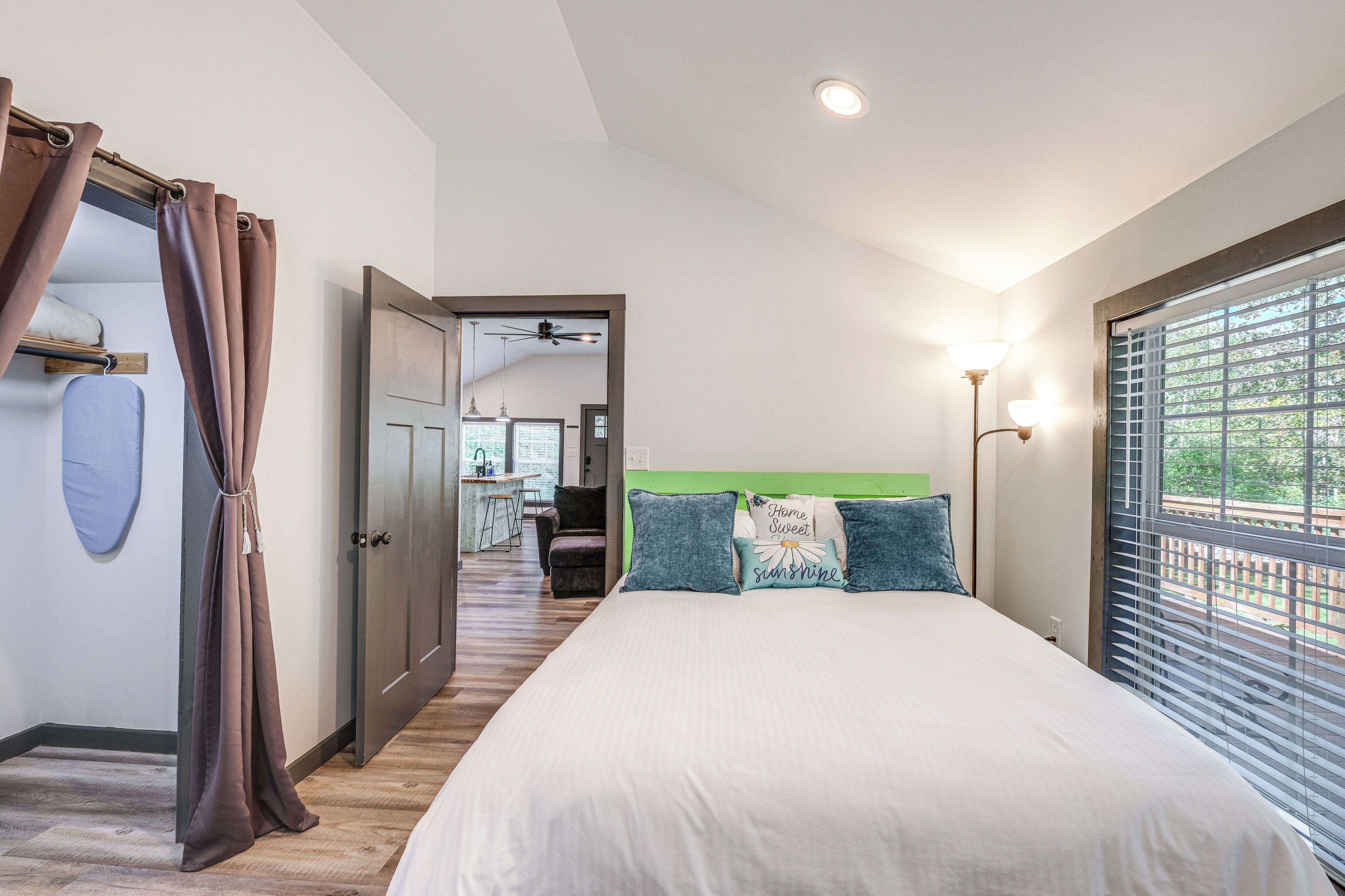 A bedroom with a large bed, a view of the outdoors through the window, a ceiling fan, and a desk with a chair in the background.