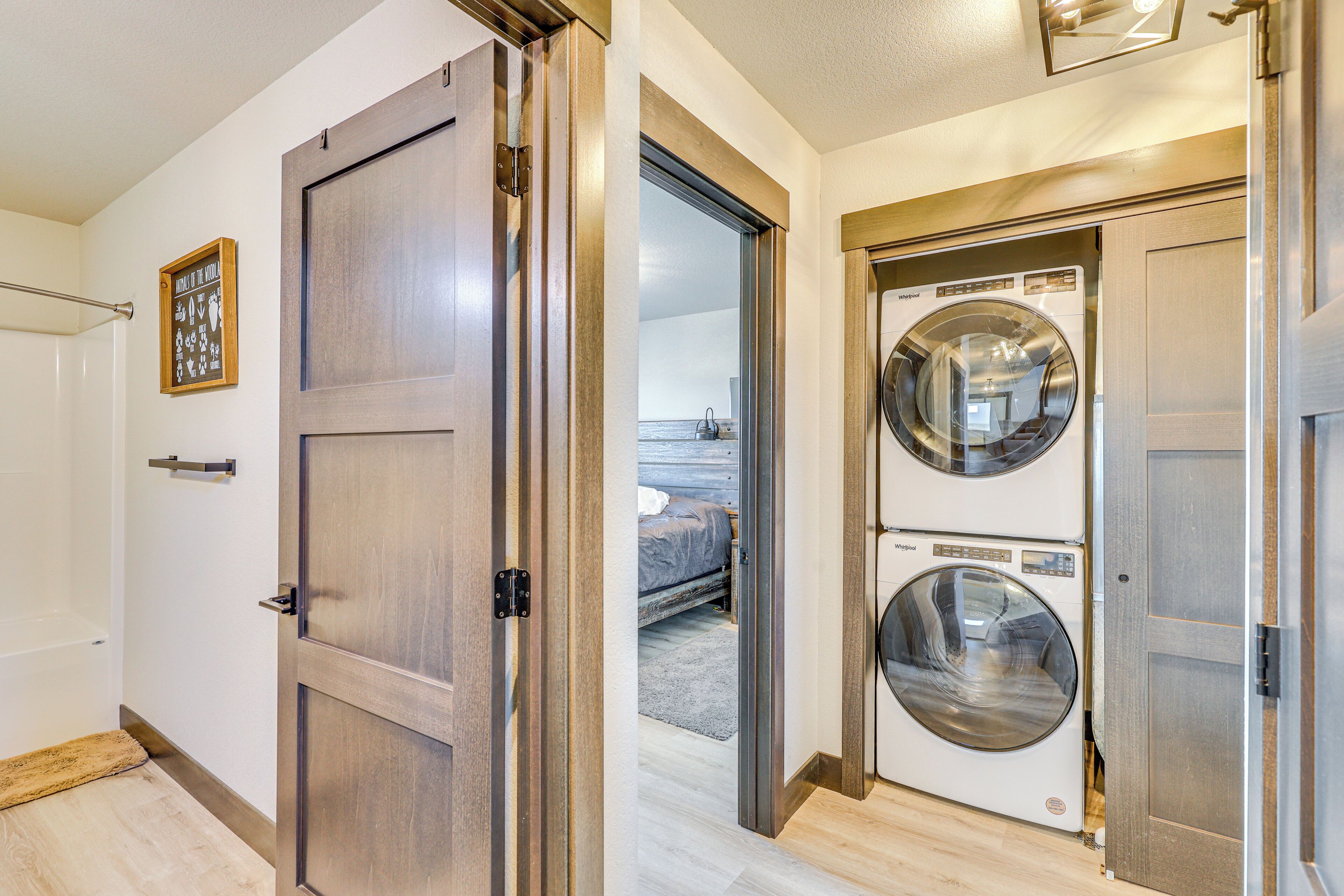 A laundry room with two stacked front-loading washing machines and a door leading to a bedroom with a bed and a nightstand.