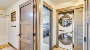 A laundry room with two stacked front-loading washing machines and a door leading to a bedroom with a bed and a nightstand.