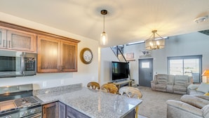 Una cocina moderna con encimeras de granito, armarios de madera y electrodomésticos de acero inoxidable. Hay una zona de comedor con una mesa y sillas, y una sala de estar con sofá y televisor.