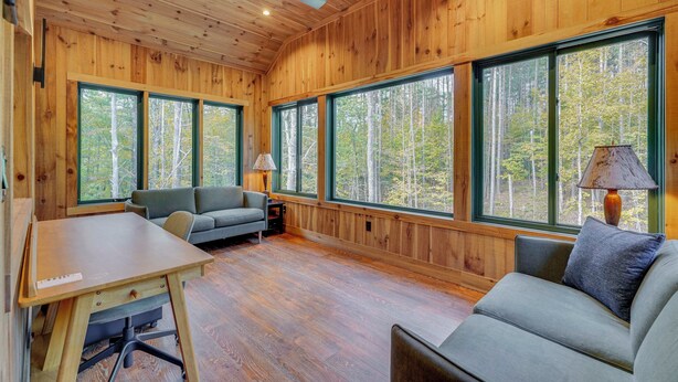 A cabin interior with wooden walls, large windows, a sofa, a table, and a lamp. - Riverfront Hideaway in Keene: Walk to Town! (Keene)