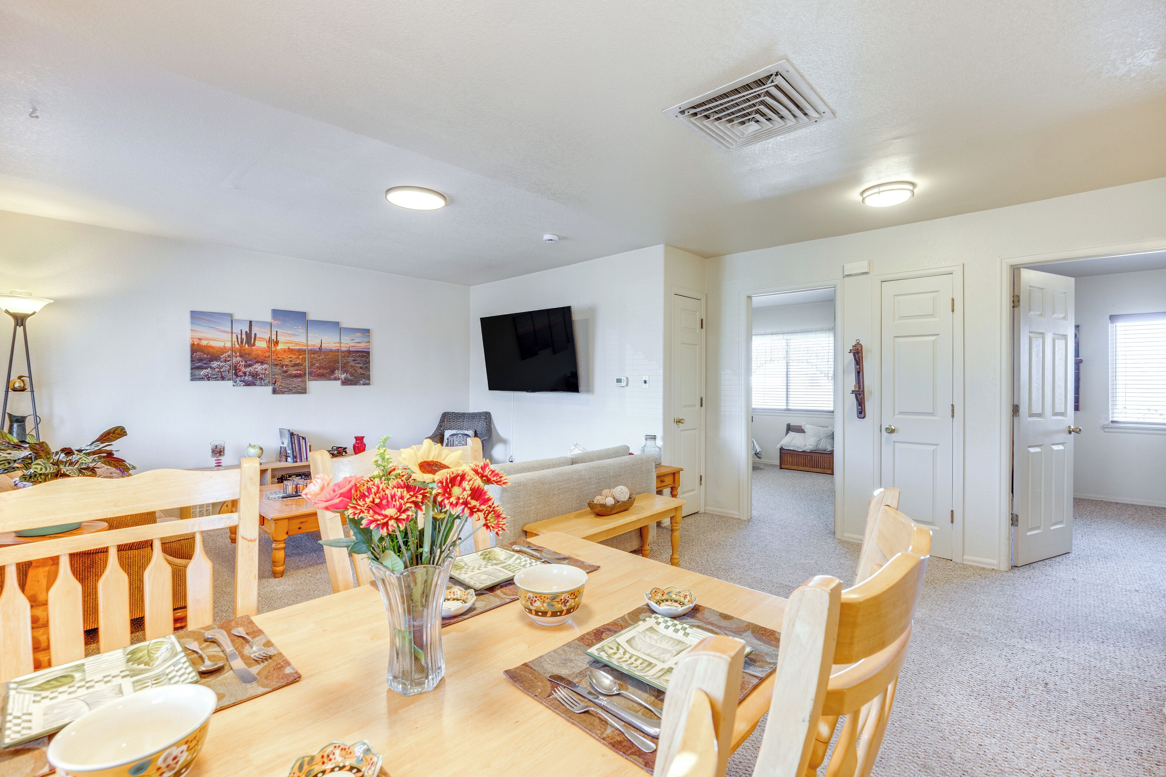 A dining area with a wooden table set for four, a vase with flowers, and a flat-screen TV mounted on the wall.