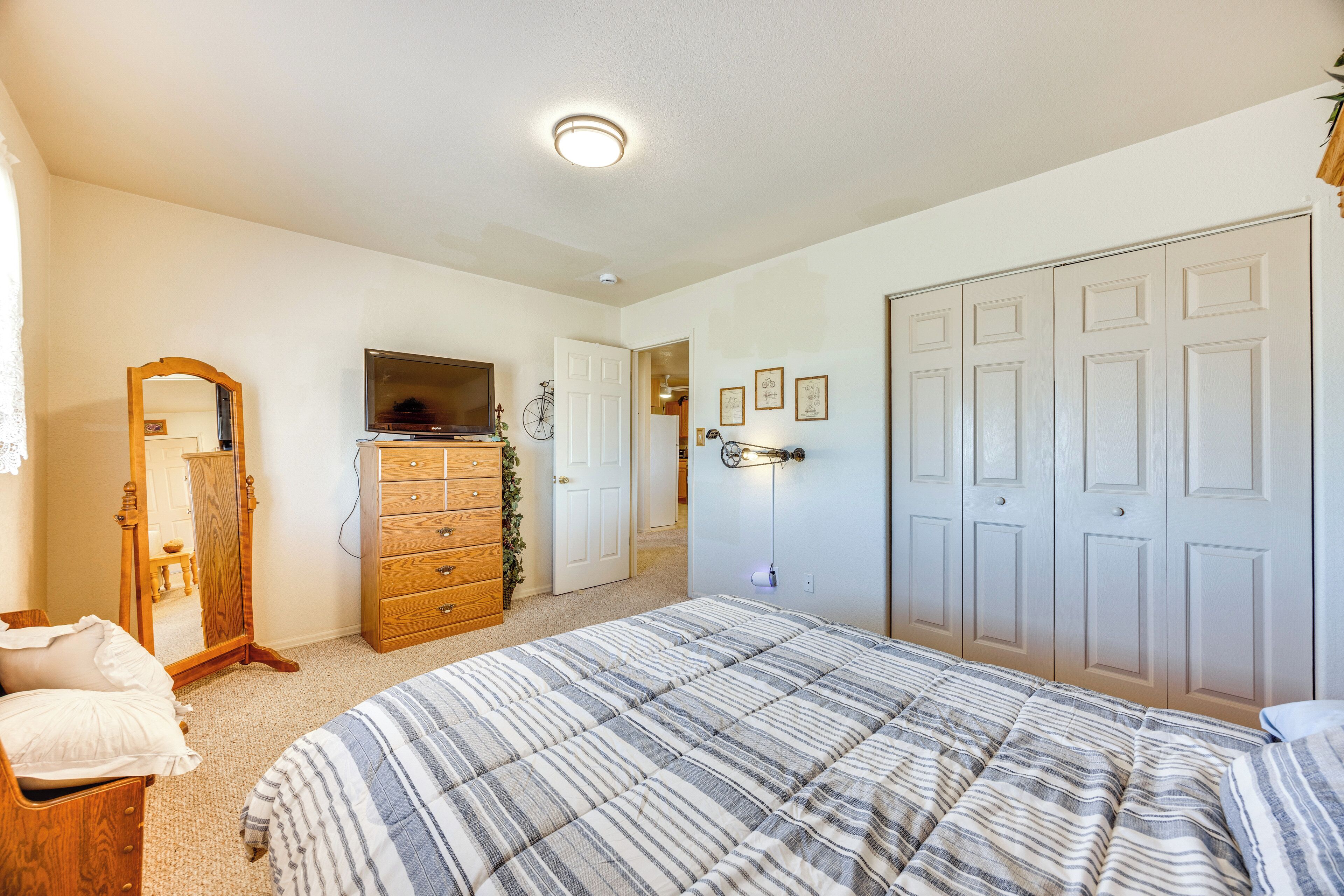 A bedroom with a large bed, wooden dresser, and a mirror. There is a door leading to another room and a wall-mounted light fixture.