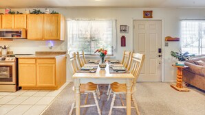 A dining area with a wooden table set for six, surrounded by wooden chairs. A kitchen with wooden cabinets and a microwave is visible to the left. - Mtn Views & Private Courtyard: Home in Pueblo West (Pueblo)
