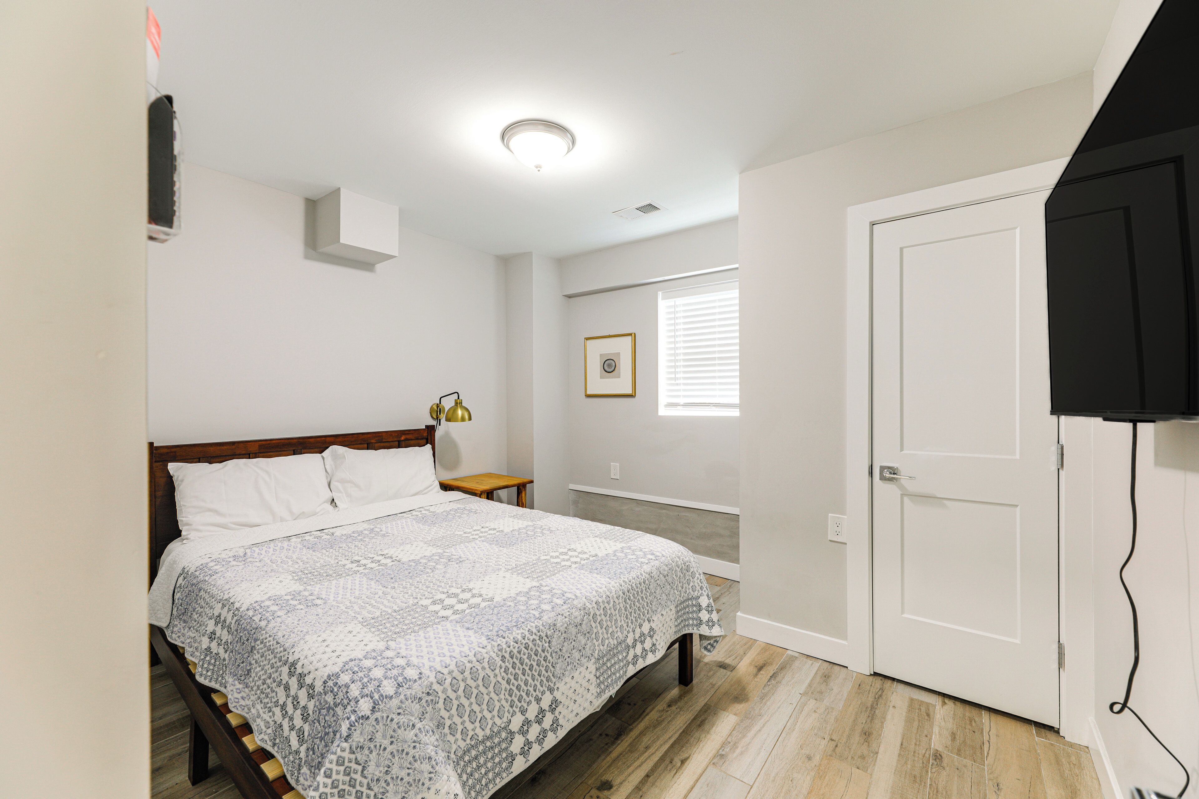 A bedroom with a bed, a bedside table with a lamp, a door, and a wall-mounted TV.