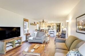 Un vaste salon doté d’un téléviseur à écran plat, d’un meuble de rangement en bois et d’une table basse. Un canapé gris et un canapé beige sont placés face au téléviseur. On aperçoit au fond une cuisine avec des meubles en bois et une table à manger.