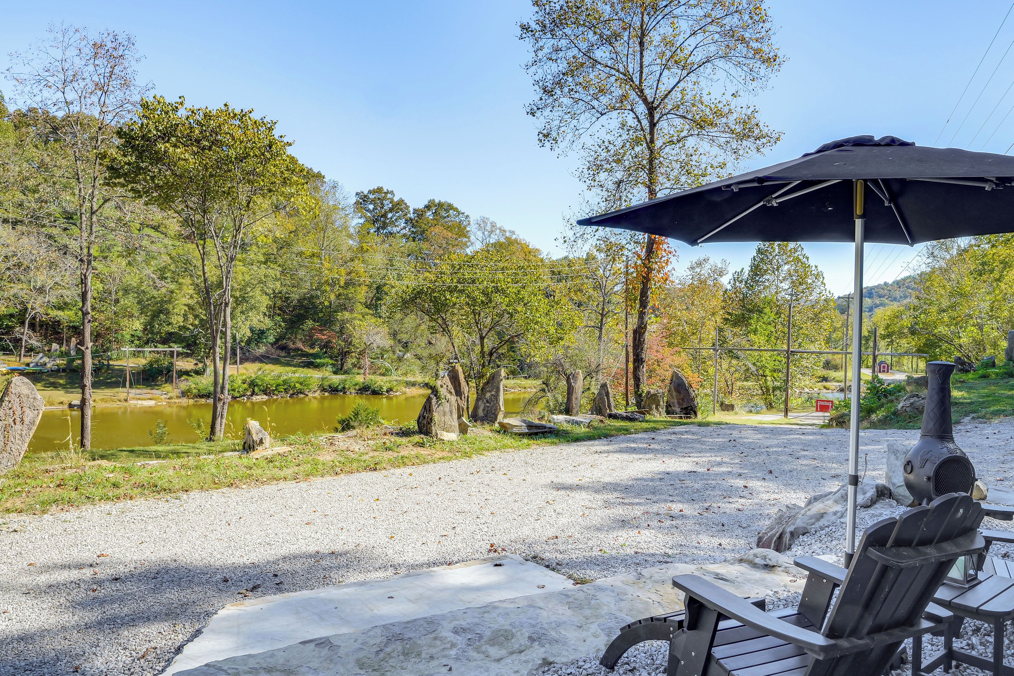 An outdoor patio with a grill, chairs, and a table under a large umbrella, surrounded by trees and a pond.