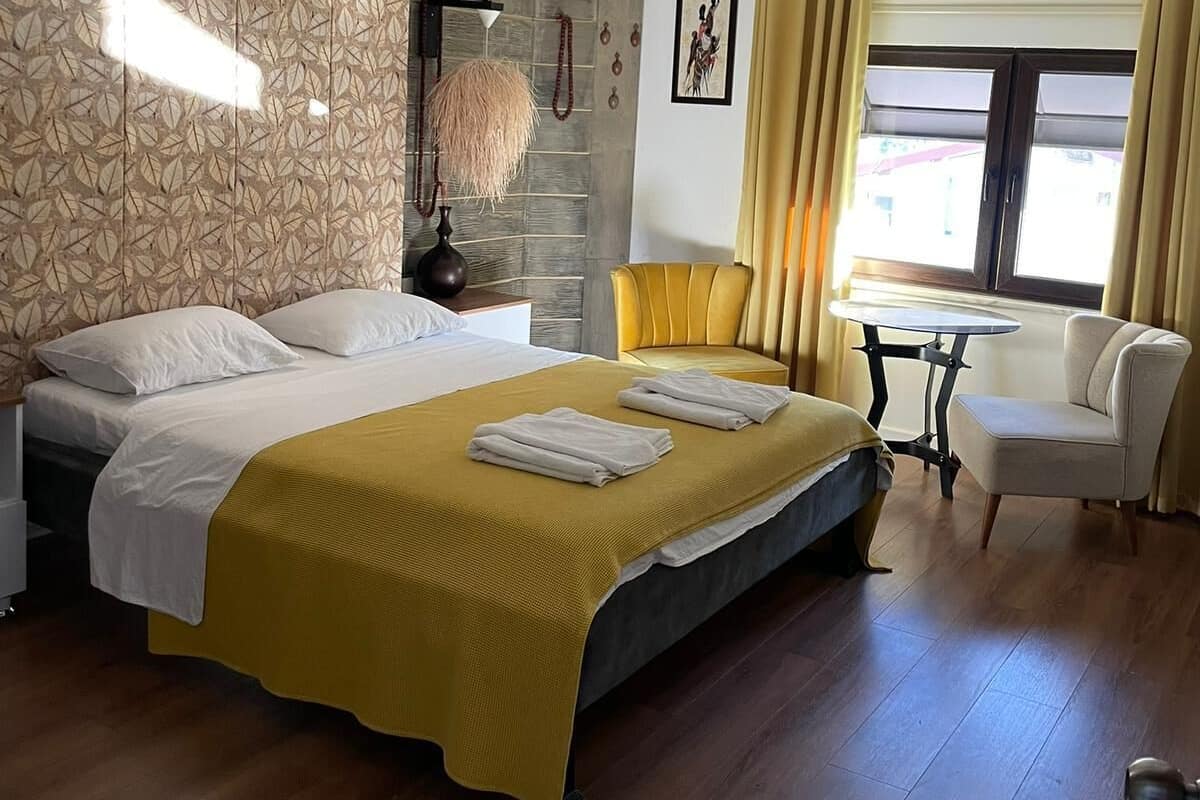 Una habitación de hotel moderna con una cama grande, una mesita, una silla y un ventanal con cortinas.