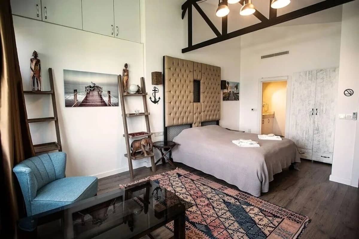 Un dormitorio con una cama grande, una silla azul, una escalera de madera, una alfombra y obras de arte en las paredes.