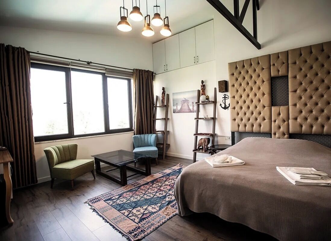 A modern bedroom with a large bed, a desk, a chair, and a sofa. There are windows with curtains, a rug, and a wall-mounted shelf with decorative items.