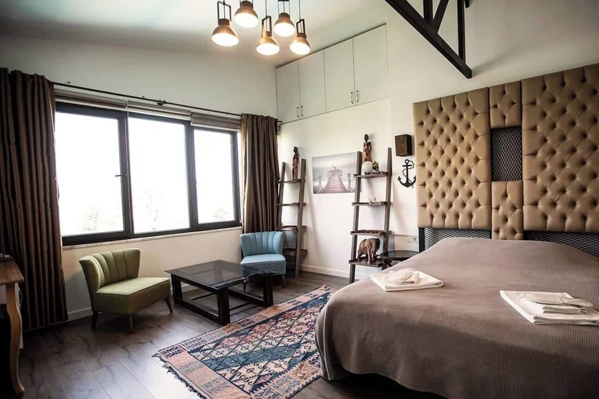 Un dormitorio moderno con una cama grande, un sofá, una mesita de centro y un escritorio. Hay ventanas con cortinas, una araña y un panel decorativo en la pared.