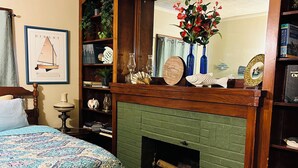 Une chambre avec une cheminée en briques vertes, un lit recouvert d’une couvre-lit à motifs, une bibliothèque et un tableau encadré au mur.