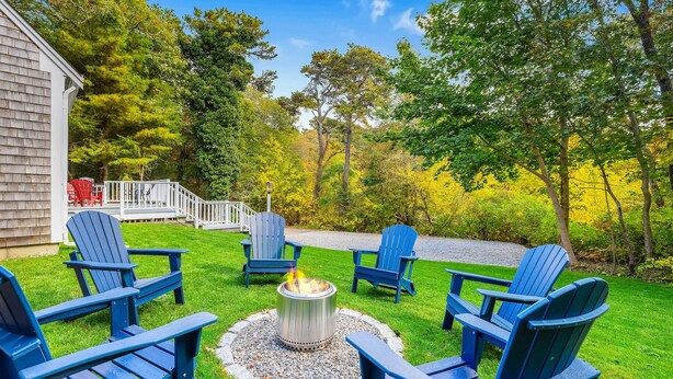 Ein Garten mit einem Feuerkorb, umgeben von blauen Adirondack-Stühlen, einer weißen Terrasse und einem Haus.