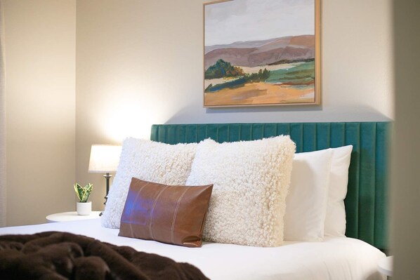 A hotel room with a bed, a headboard, a painting, a nightstand, a lamp, and a plant.