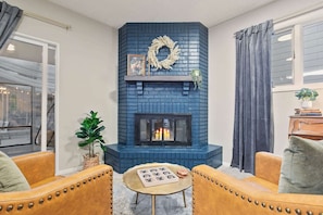 Un salon avec une cheminée en briques bleues, des fauteuils en cuir, une petite table ronde et une vue sur la rue.
