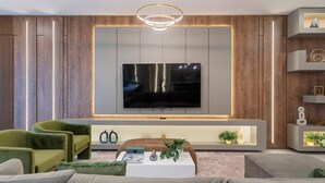 A modern living room with a large flat-screen TV mounted on a wooden panel wall, a white console table with decorative items, and green upholstered chairs. - 5911 Luxury 10bd Family Home For an Unforgettable Vacation (Orlando)