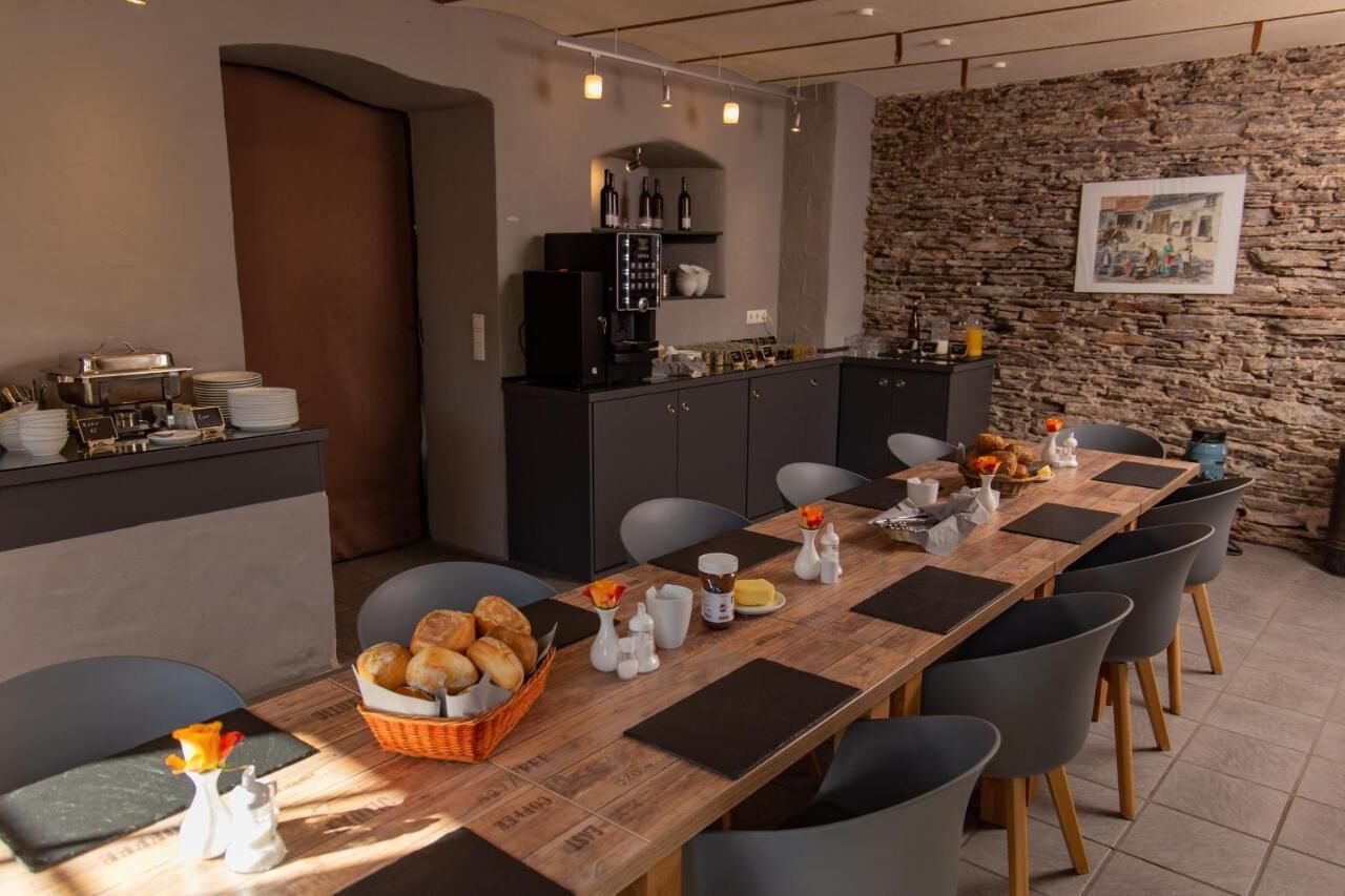 A dining area with a long wooden table set for multiple people, surrounded by chairs. There is a buffet counter with various dishes and a coffee machine. The room features a stone wall and a framed picture on the wall.