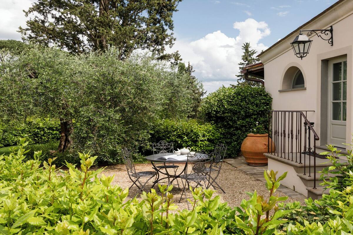 A patio with a table and chairs, surrounded by lush greenery and a building with a balcony.