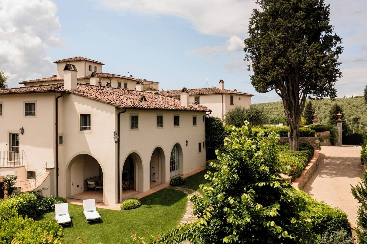 A large, two-story building with a tiled roof, multiple windows, and a well-maintained garden with a lawn and a few trees.