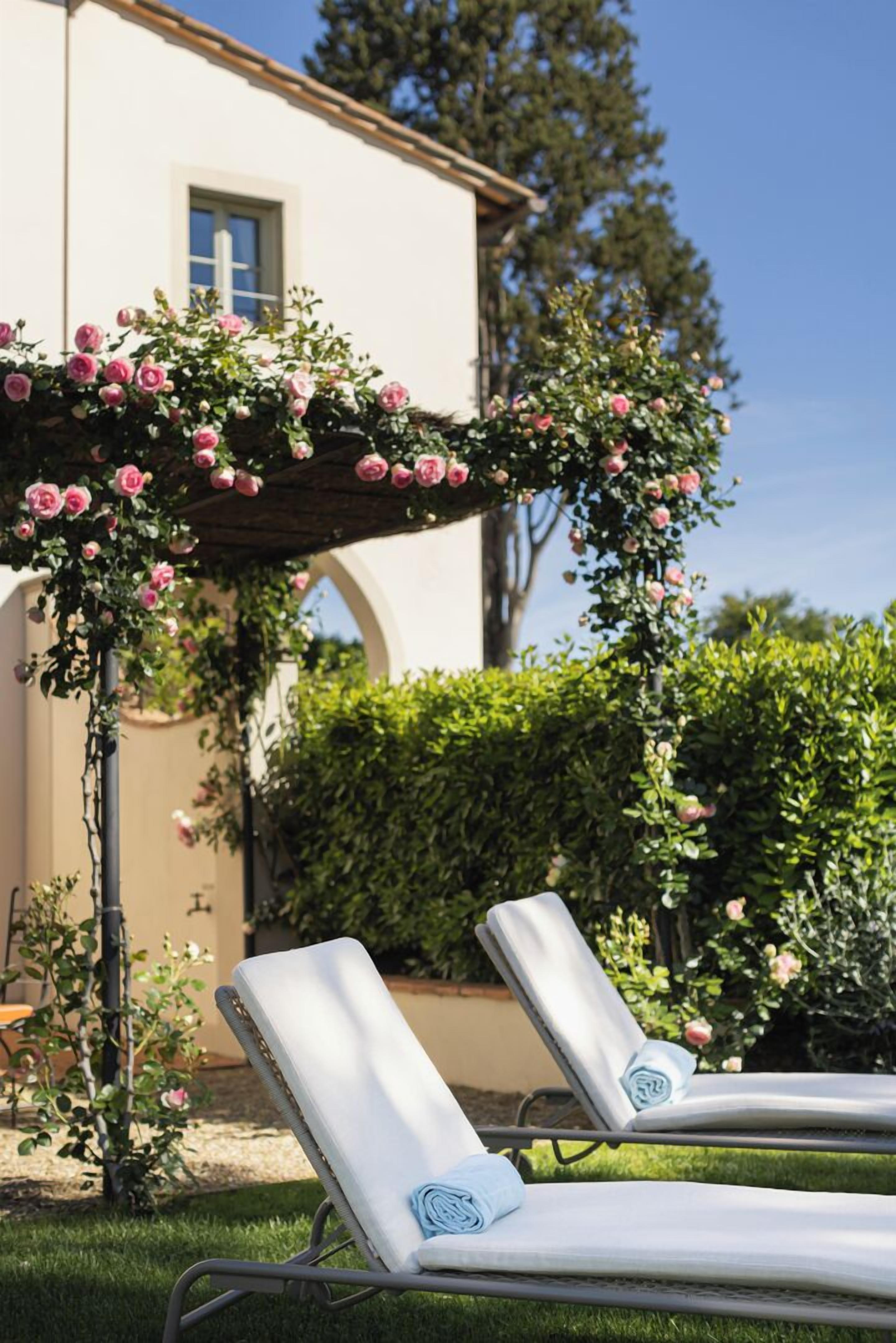 Ein Garten mit einer Pergola, die in pinkfarbene Rosen bewachsen ist, zwei weiße Liegestühle mit Handtüchern und ein Gebäude mit heller Fassade und Bogenfenstern.