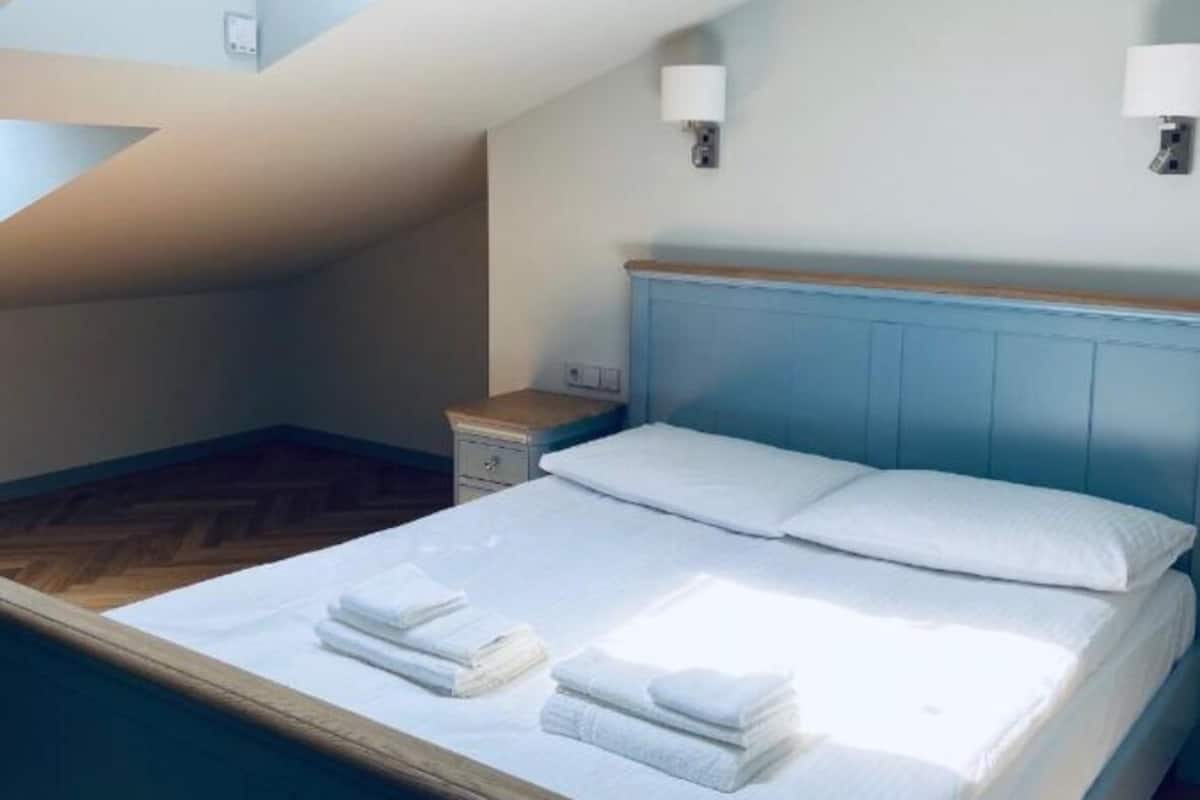 A bedroom with a sloped ceiling, a bed with white linens, two wall-mounted lamps, and a wooden nightstand.