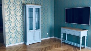 A room with a herringbone wooden floor, a light blue patterned wall, a white cabinet with glass doors, and a white TV stand.