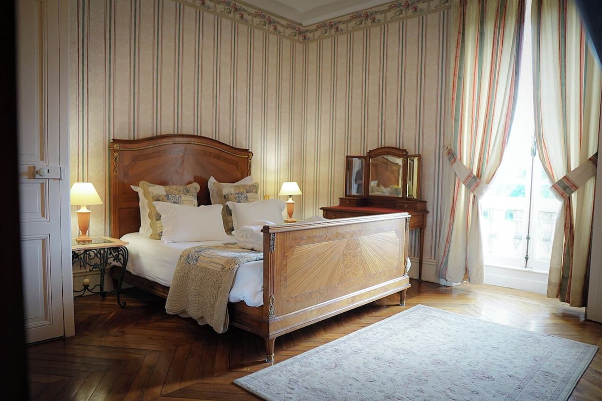 A bedroom with a wooden bed, two bedside tables with lamps, a dresser, and a large window with curtains.