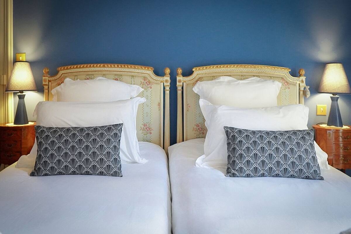 A double bed with white linens and patterned pillows, flanked by two bedside tables with lamps, set against a blue wall with floral wallpaper.
