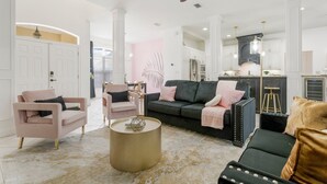 A spacious living room with a dark green sofa, a pink armchair, and a black leather sofa. There is a round coffee table with a decorative object on it. The room has white walls and a ceiling fan.