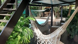 Eine Hängematte unter einem schattigen Bereich mit Pool und Gartenmöbeln, umgeben von üppigem Grün.