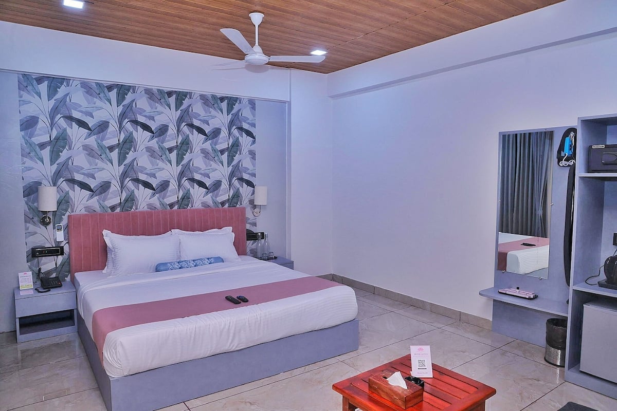 A modern hotel room with a large bed, a ceiling fan, a nightstand with a phone, a bedside table with a lamp, and a small seating area with a table and chairs.