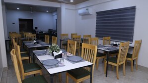 A dining area with wooden chairs and tables, an air conditioning unit, and a window with blinds. - Hotel Lotus (Botad)