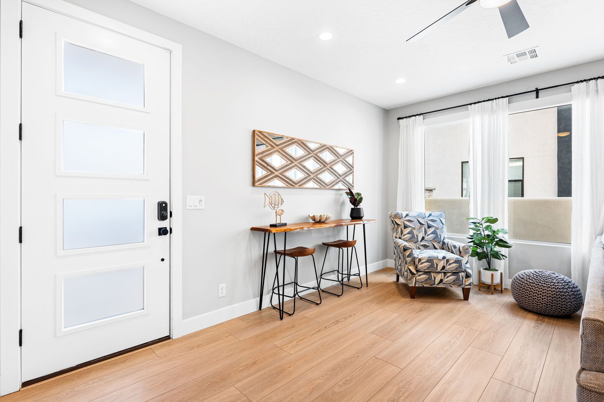 Ein modernes Wohnzimmer mit Holzboden, einer weißen Tür, einem Essbereich mit Hockern, einem gemusterten Sessel und einem großen Fenster mit Vorhängen.